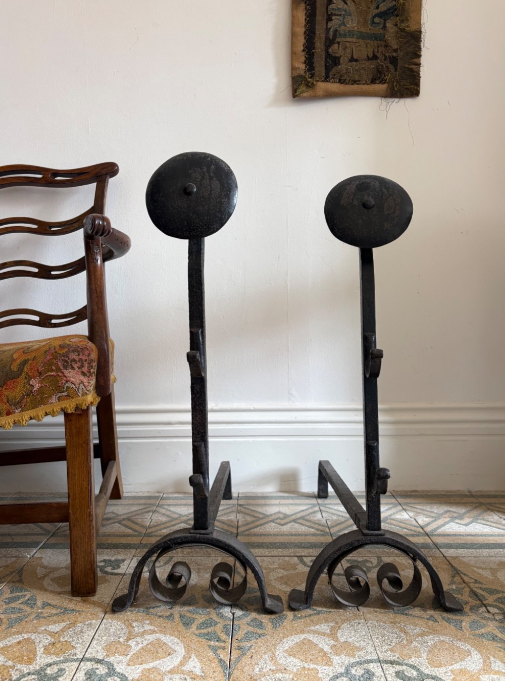 A pair of hand-forged black wrought iron fire dogs featuring circular disc tops, tool hooks, and scrolled arched feet. View II