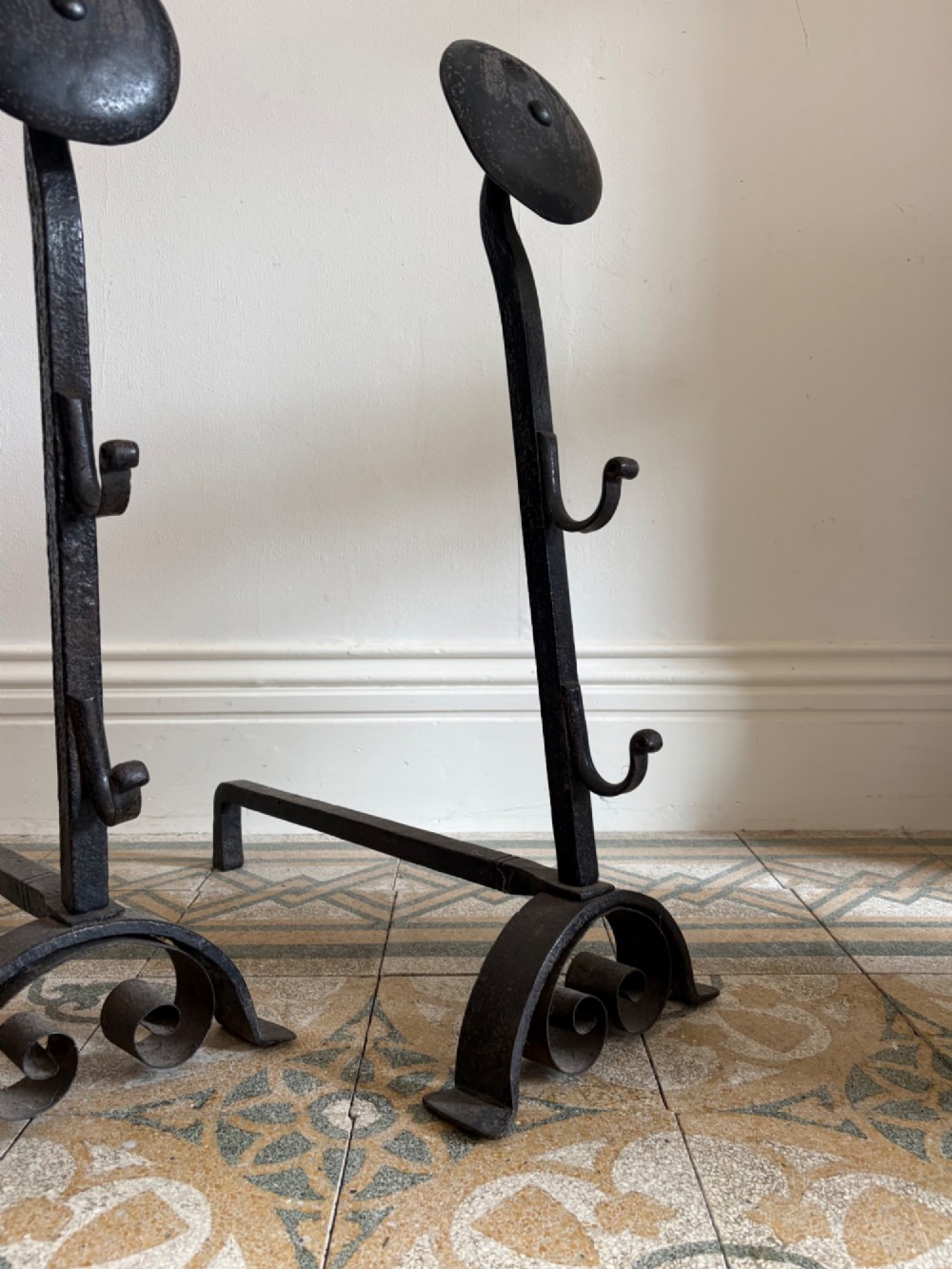 A pair of hand-forged black wrought iron fire dogs featuring circular disc tops, tool hooks, and scrolled arched feet. View V