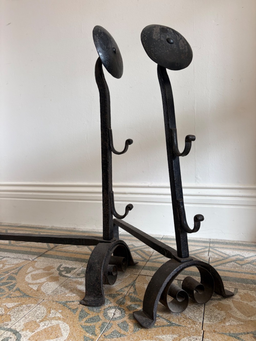 A pair of hand-forged black wrought iron fire dogs featuring circular disc tops, tool hooks, and scrolled arched feet. View VIII