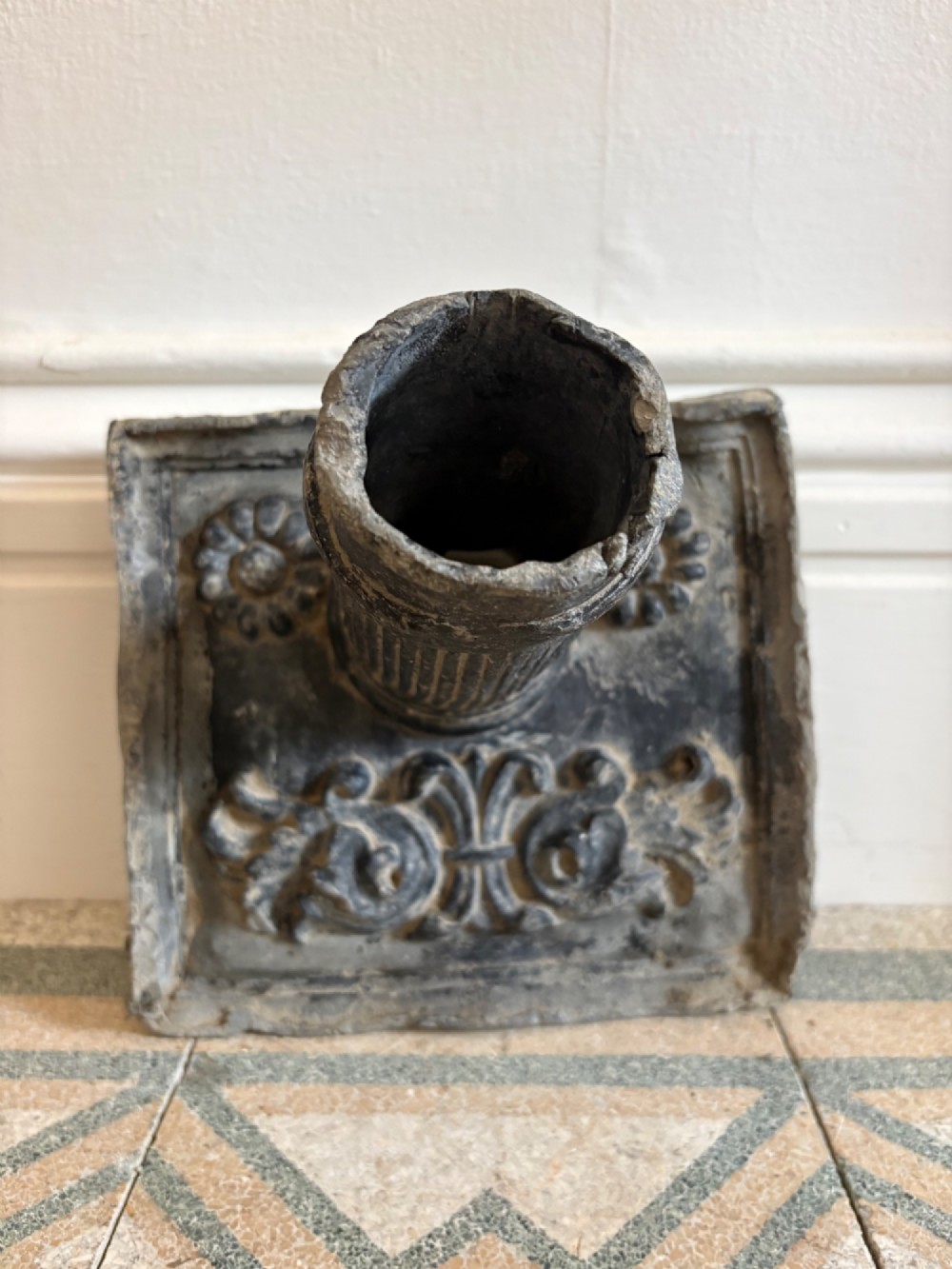 A square dark grey cast lead fountain head featuring a central cylindrical spout above relief-moulded floral rosettes and scrollwork motifs. View II
