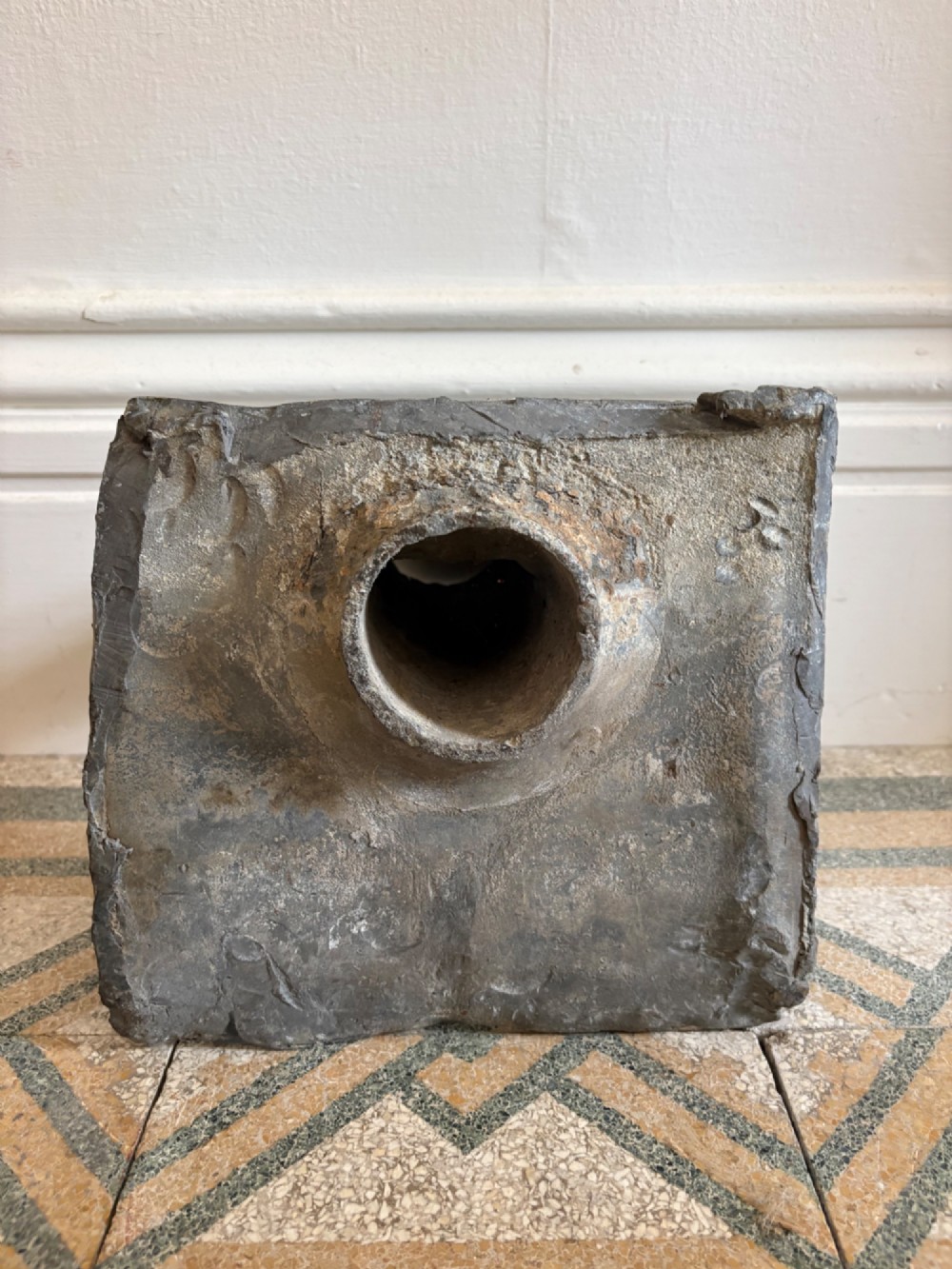 A square dark grey cast lead fountain head featuring a central cylindrical spout above relief-moulded floral rosettes and scrollwork motifs. View III