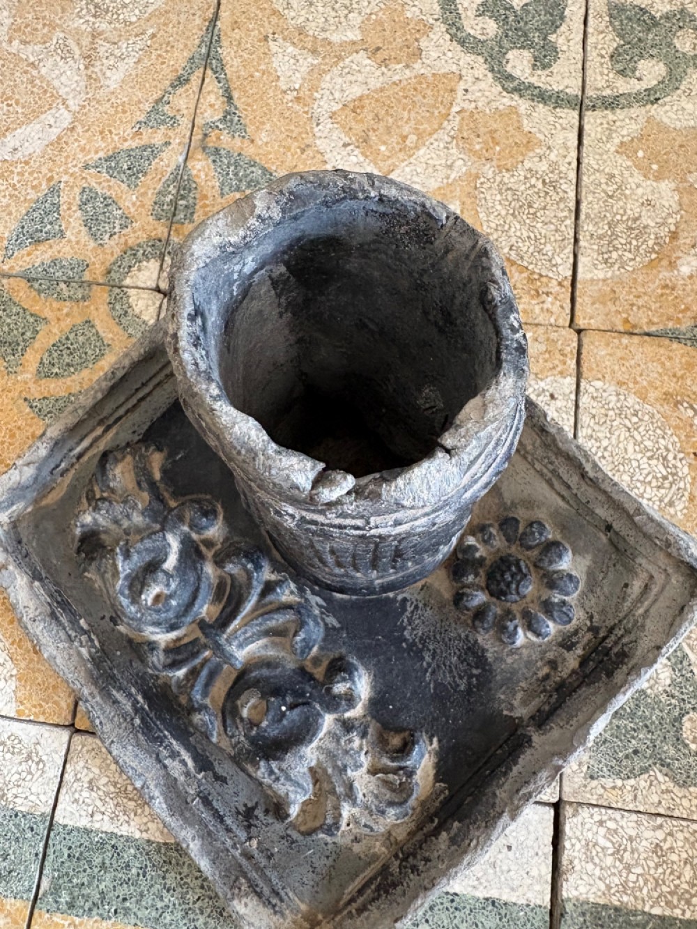 A square dark grey cast lead fountain head featuring a central cylindrical spout above relief-moulded floral rosettes and scrollwork motifs. View V