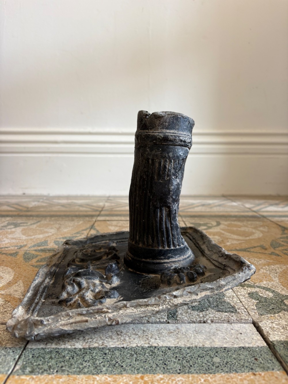 A square dark grey cast lead fountain head featuring a central cylindrical spout above relief-moulded floral rosettes and scrollwork motifs. View VI