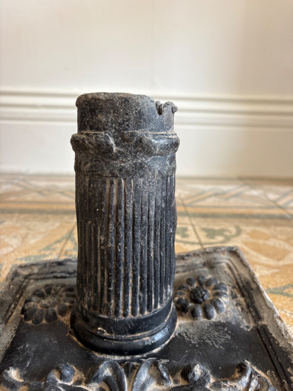 A square dark grey cast lead fountain head featuring a central cylindrical spout above relief-moulded floral rosettes and scrollwork motifs. View IX