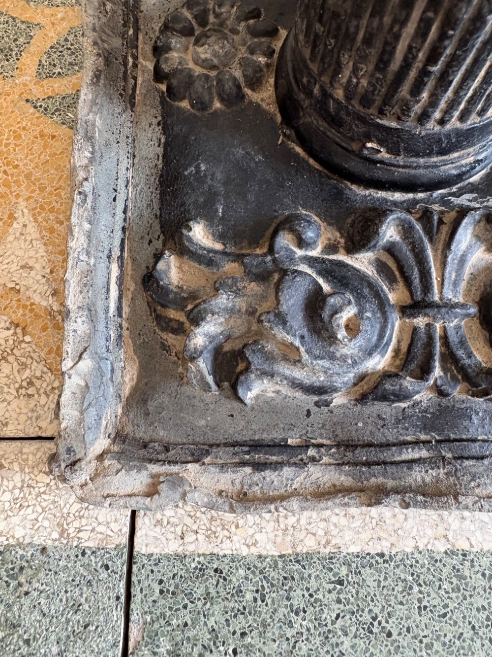 A square dark grey cast lead fountain head featuring a central cylindrical spout above relief-moulded floral rosettes and scrollwork motifs. View X