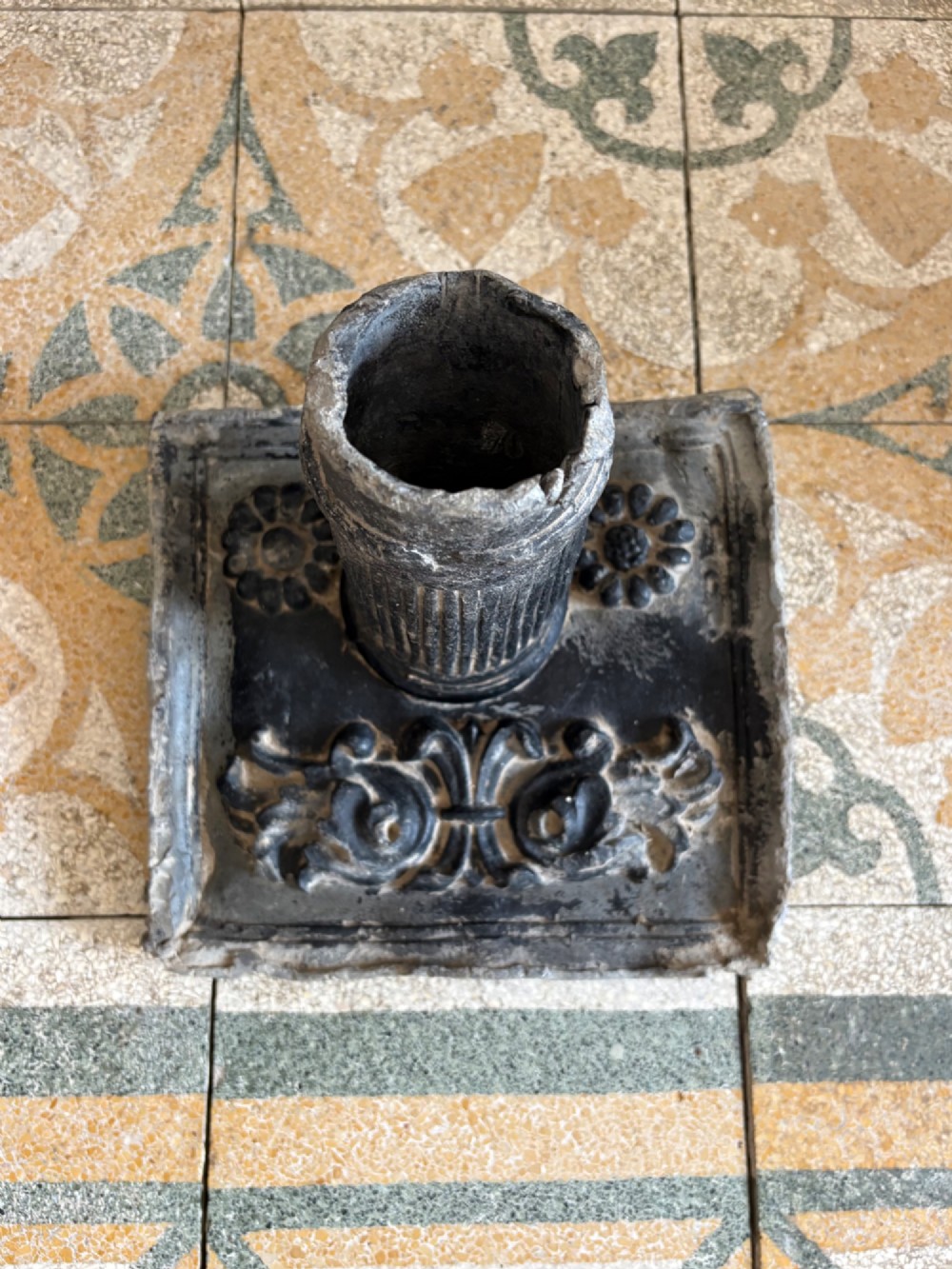 A square dark grey cast lead fountain head featuring a central cylindrical spout above relief-moulded floral rosettes and scrollwork motifs. View XII