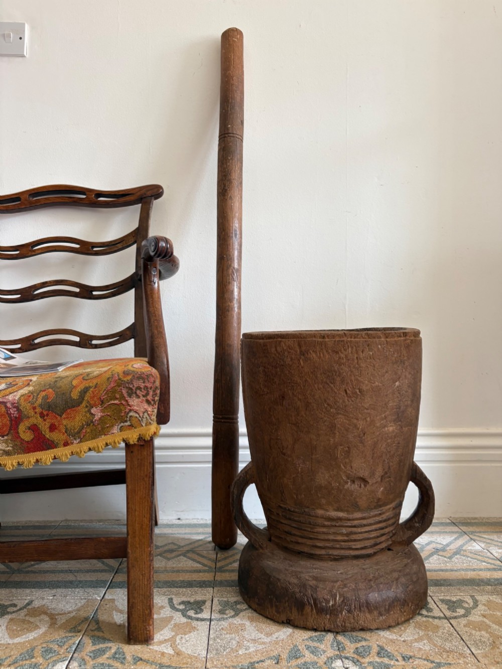 A hand-carved African hardwood grain mortar with side loop handles and concentric carved grooves, next to a tall wooden pestle pole.