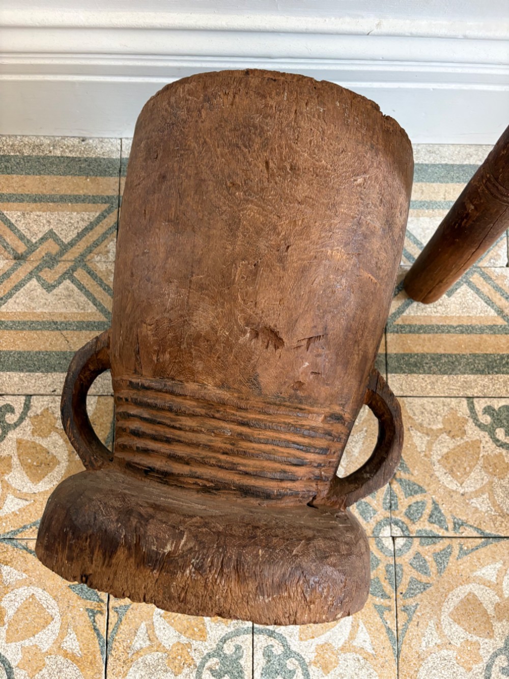A hand-carved African hardwood grain mortar with side loop handles and concentric carved grooves, next to a tall wooden pestle pole. View III