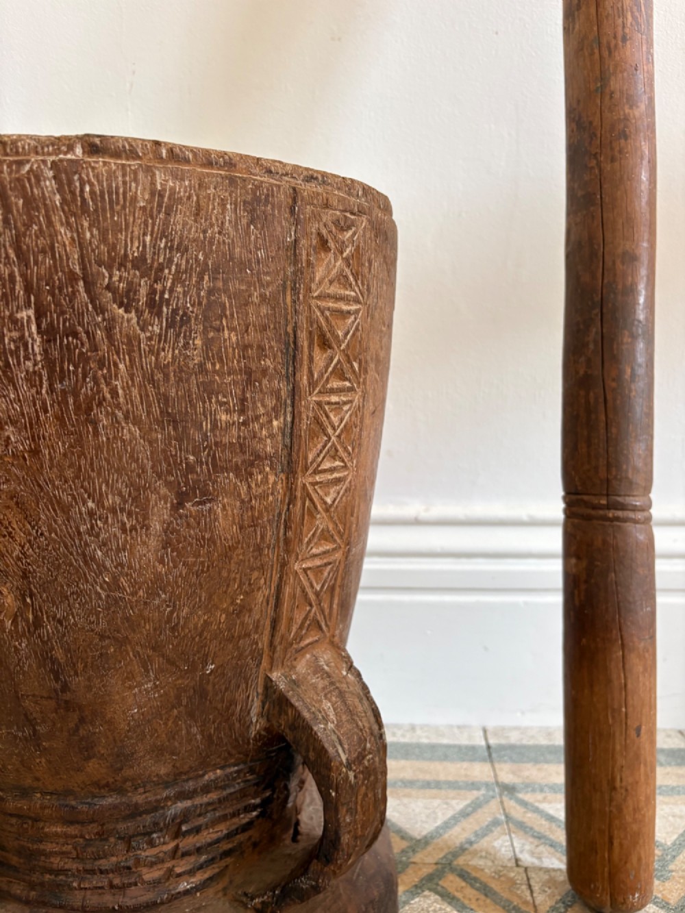 A hand-carved African hardwood grain mortar with side loop handles and concentric carved grooves, next to a tall wooden pestle pole. View V