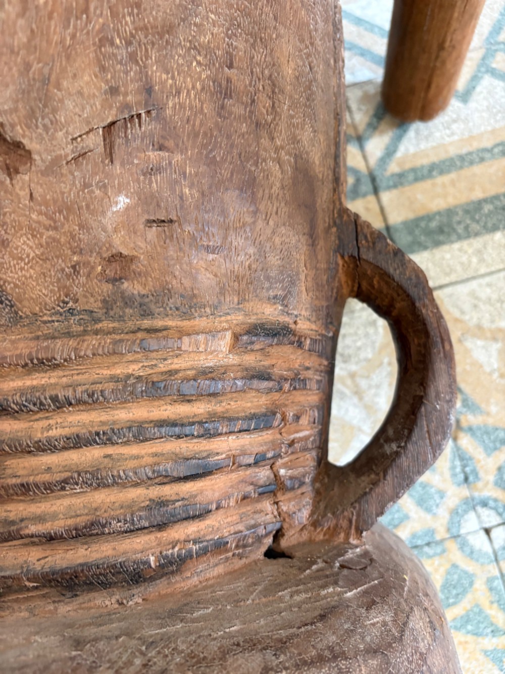 A hand-carved African hardwood grain mortar with side loop handles and concentric carved grooves, next to a tall wooden pestle pole. View VII