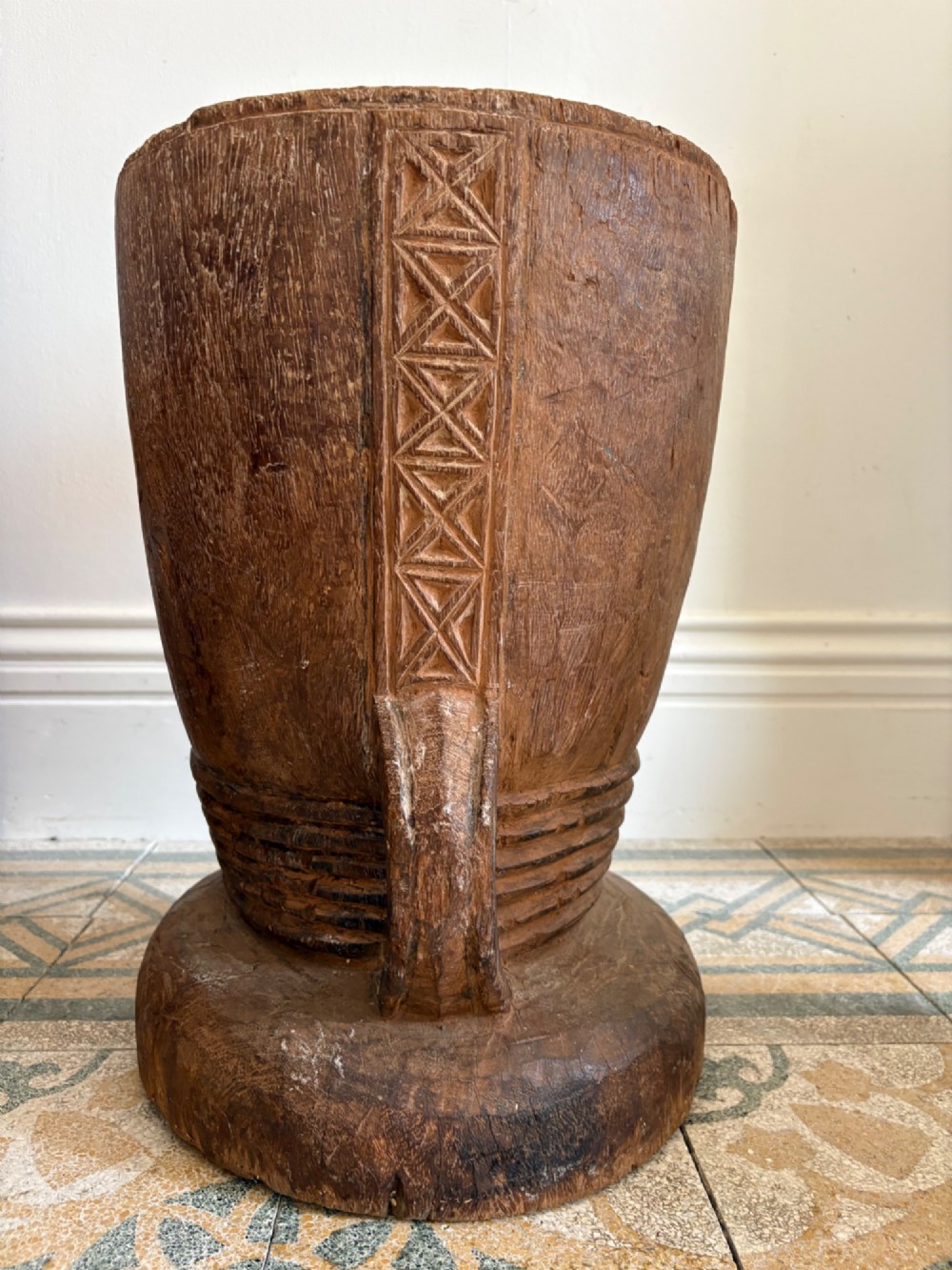 A hand-carved African hardwood grain mortar with side loop handles and concentric carved grooves, next to a tall wooden pestle pole. View IX