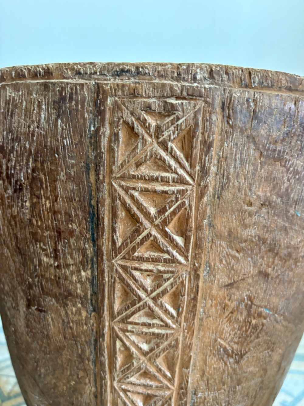A hand-carved African hardwood grain mortar with side loop handles and concentric carved grooves, next to a tall wooden pestle pole. View X