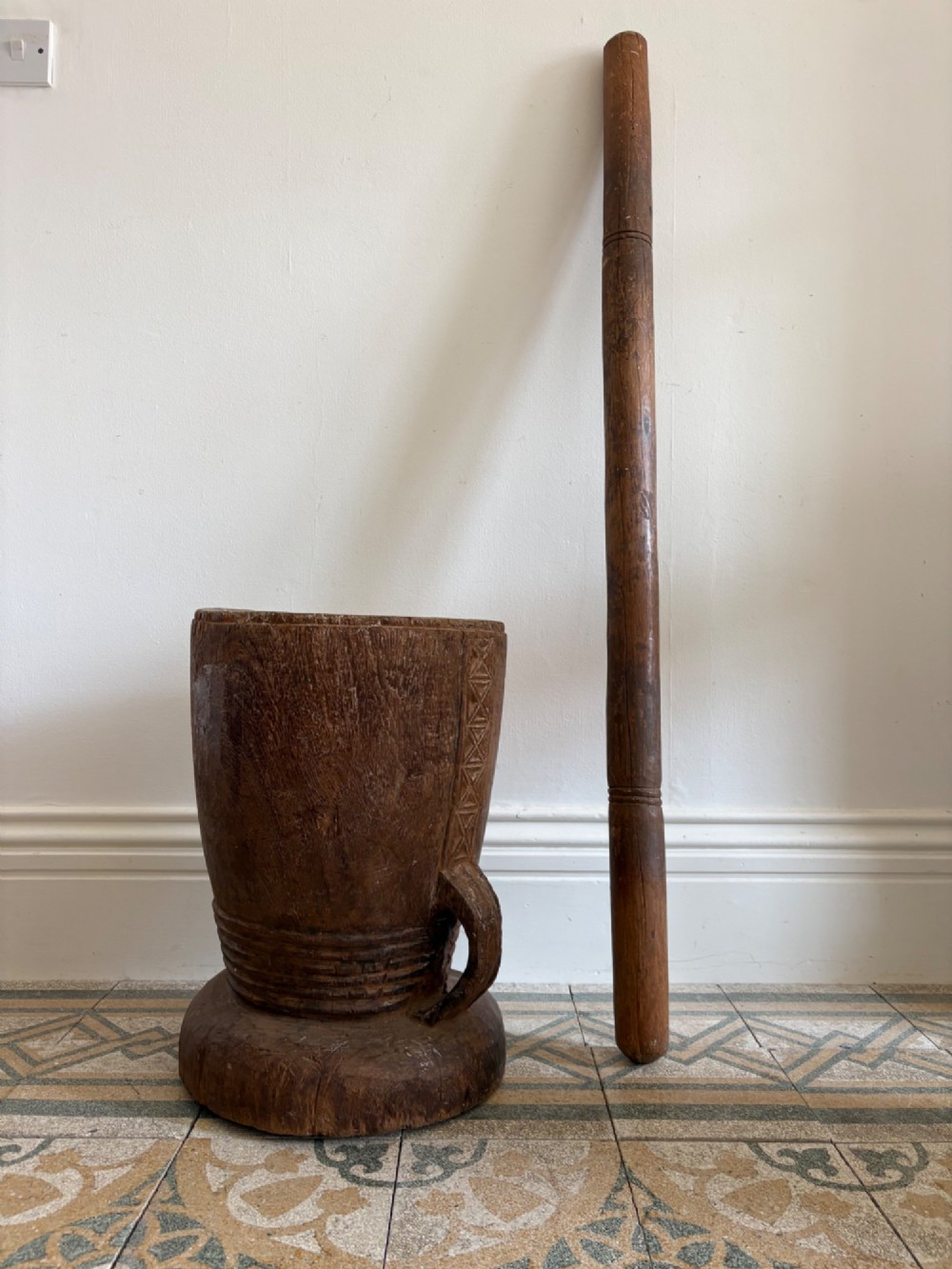 A hand-carved African hardwood grain mortar with side loop handles and concentric carved grooves, next to a tall wooden pestle pole. View XI