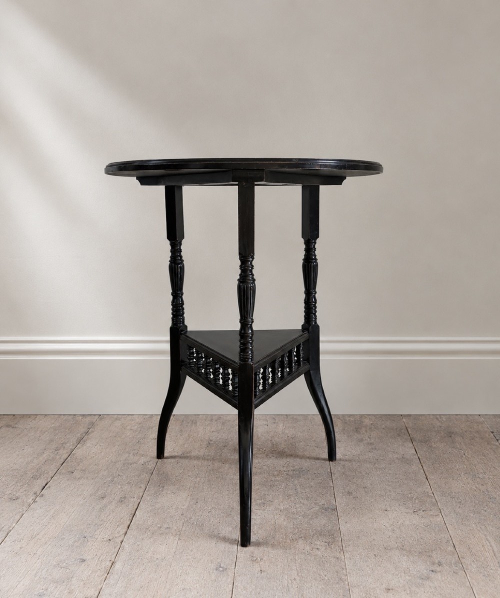 A late 19th-century ebonised round side table featuring three turned legs, slender splayed feet, and a lower triangular shelf bounded by a spindled gallery.