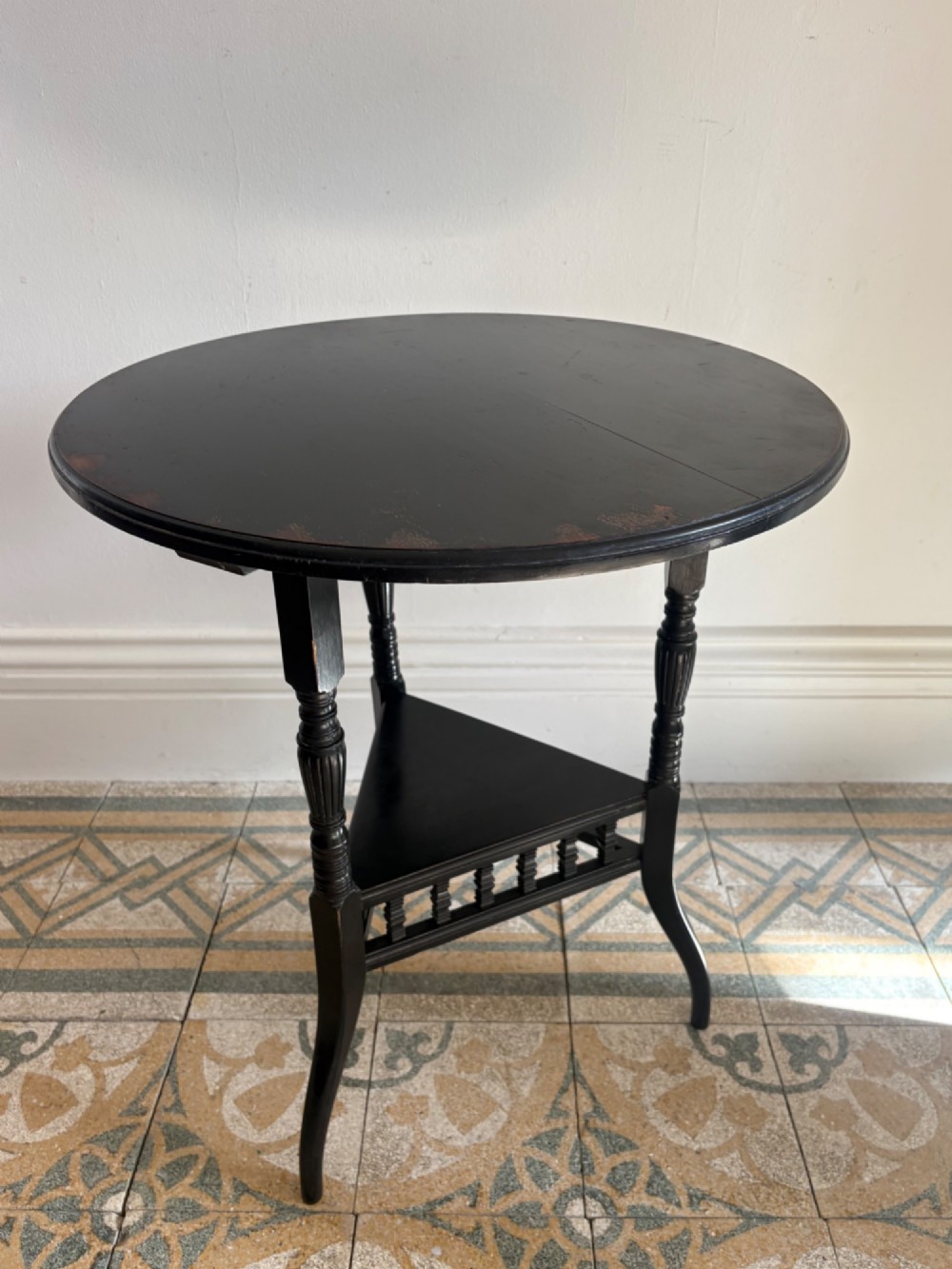 A late 19th-century ebonised round side table featuring three turned legs, slender splayed feet, and a lower triangular shelf bounded by a spindled gallery. View VII