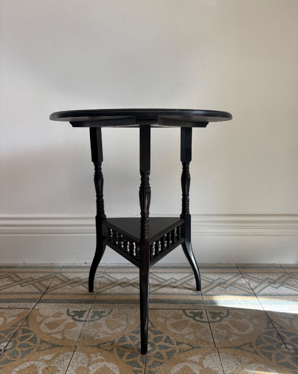 A late 19th-century ebonised round side table featuring three turned legs, slender splayed feet, and a lower triangular shelf bounded by a spindled gallery. View X