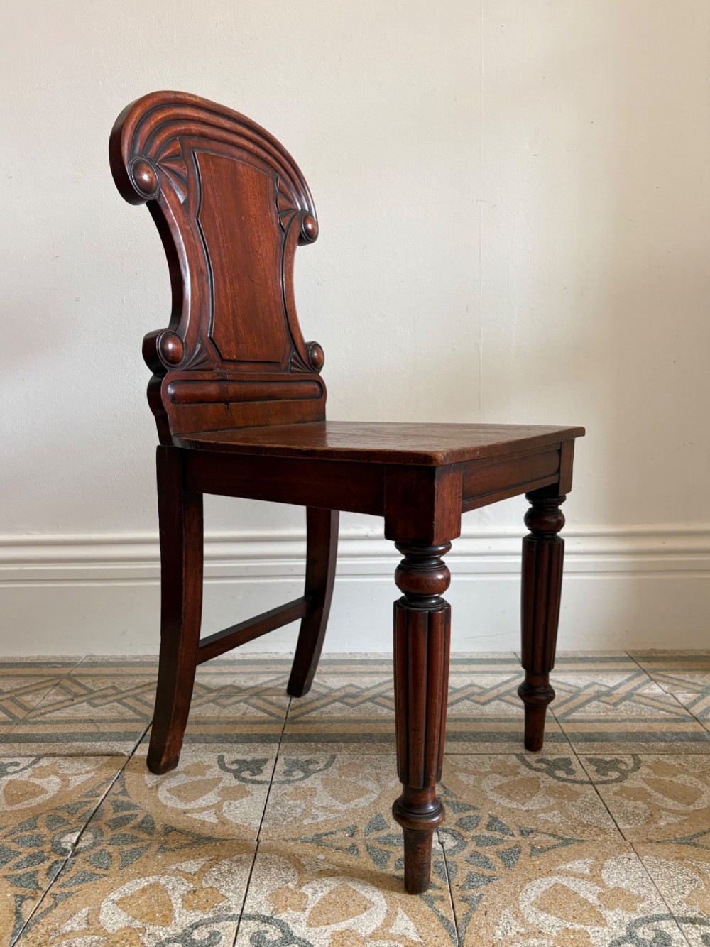 An antique William IV mahogany hall chair featuring a carved crest backrest, solid flat seat, and fluted turned front legs.