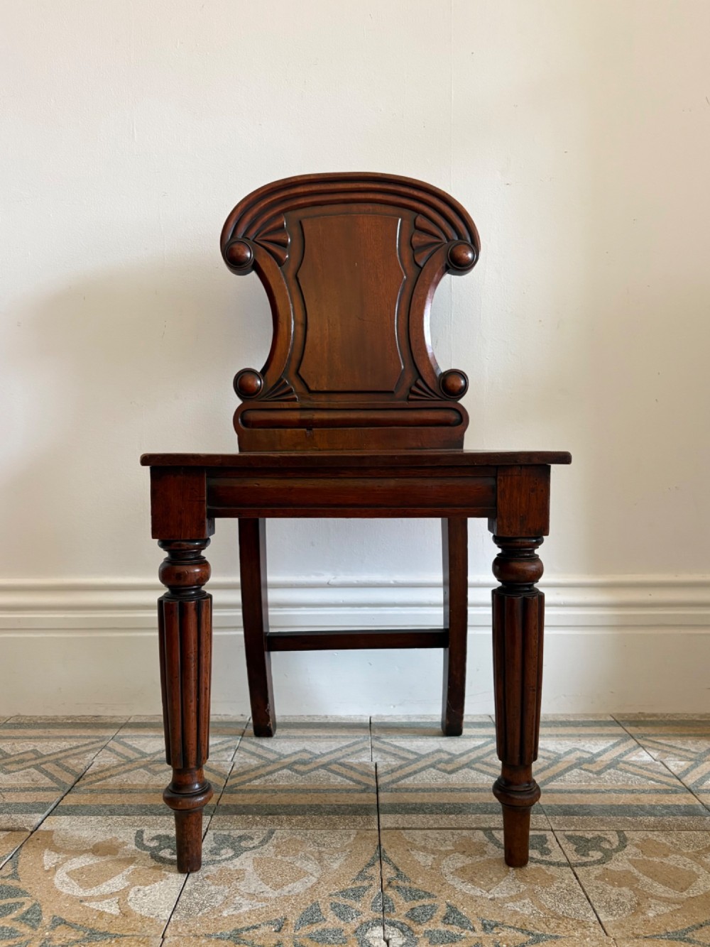 An antique William IV mahogany hall chair featuring a carved crest backrest, solid flat seat, and fluted turned front legs. View IV