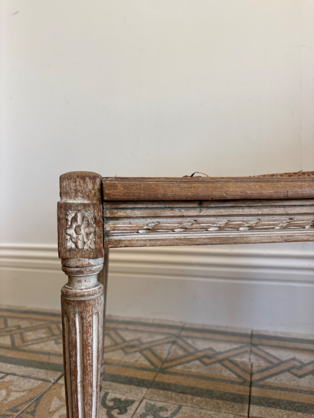 A French Neoclassical rectangular stool featuring a distressed white painted finish, fluted tapered legs, carved rosette corner details, and a woven cane seat. View II