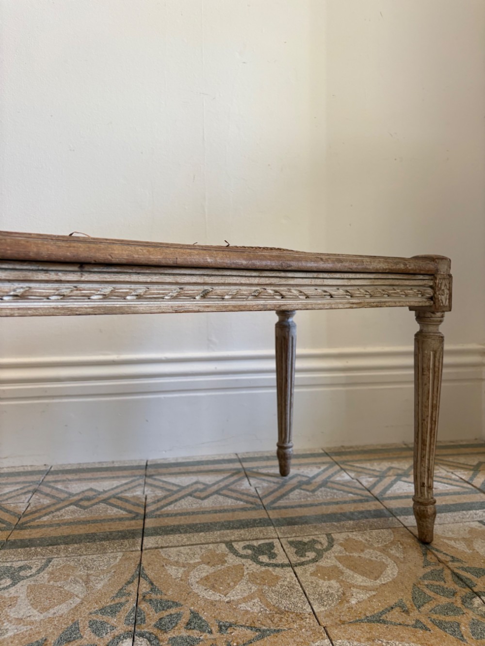 A French Neoclassical rectangular stool featuring a distressed white painted finish, fluted tapered legs, carved rosette corner details, and a woven cane seat. View III