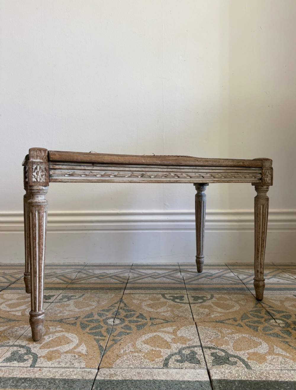 A French Neoclassical rectangular stool featuring a distressed white painted finish, fluted tapered legs, carved rosette corner details, and a woven cane seat. View VII