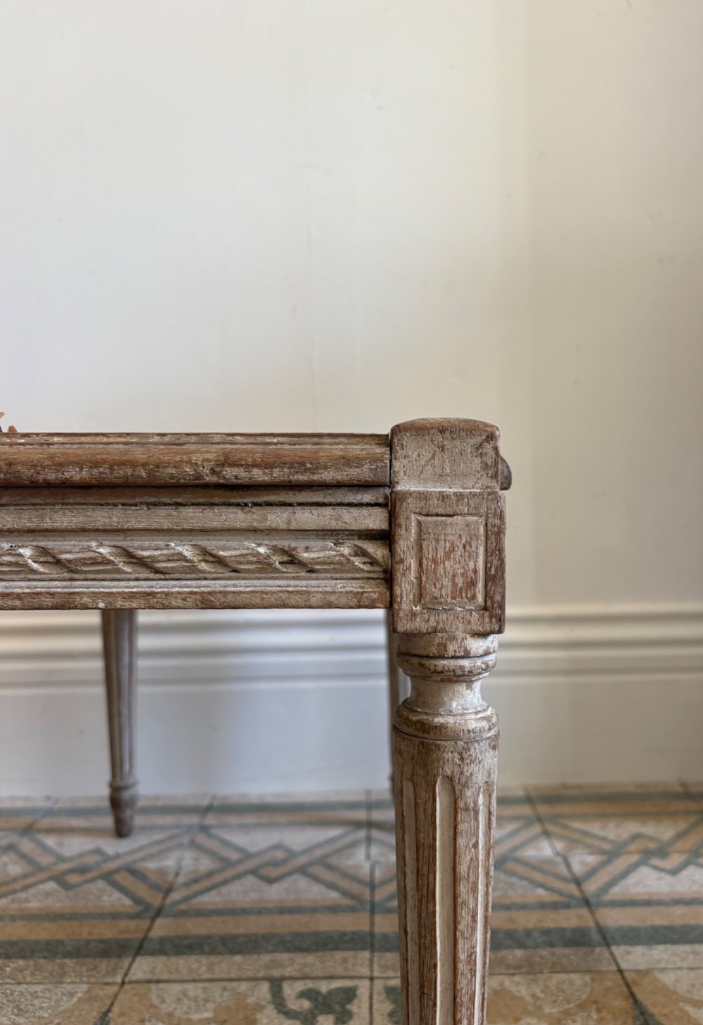 A French Neoclassical rectangular stool featuring a distressed white painted finish, fluted tapered legs, carved rosette corner details, and a woven cane seat. View VIII