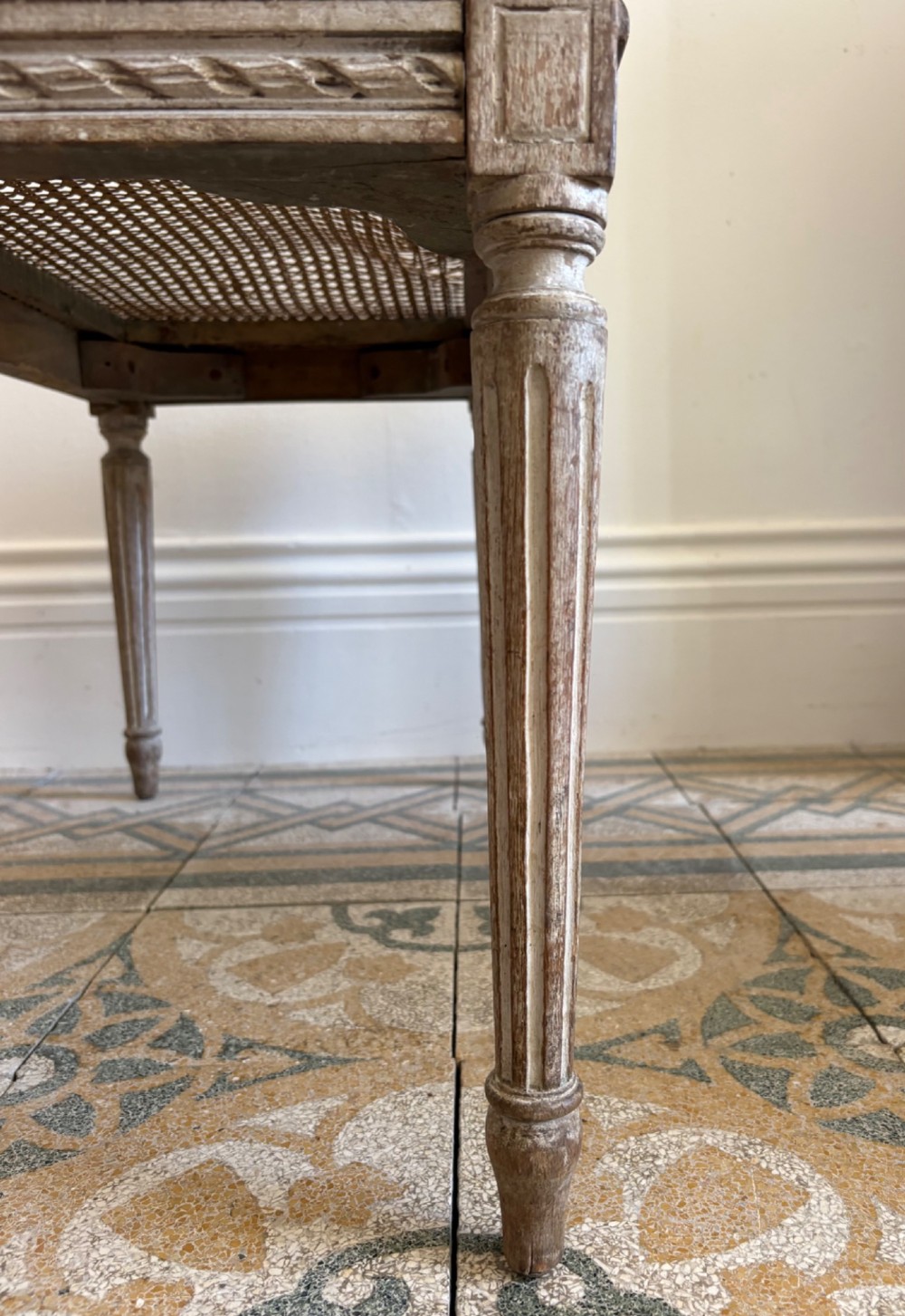 A French Neoclassical rectangular stool featuring a distressed white painted finish, fluted tapered legs, carved rosette corner details, and a woven cane seat. View IX