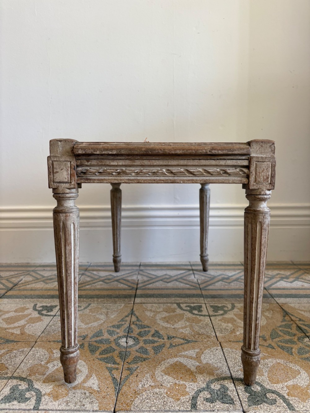 A French Neoclassical rectangular stool featuring a distressed white painted finish, fluted tapered legs, carved rosette corner details, and a woven cane seat. View X
