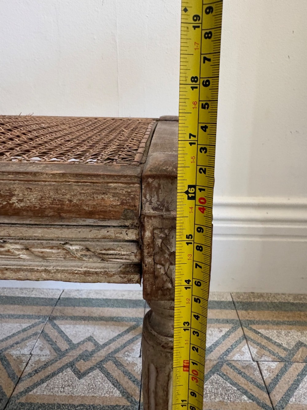 A French Neoclassical rectangular stool featuring a distressed white painted finish, fluted tapered legs, carved rosette corner details, and a woven cane seat. View XV