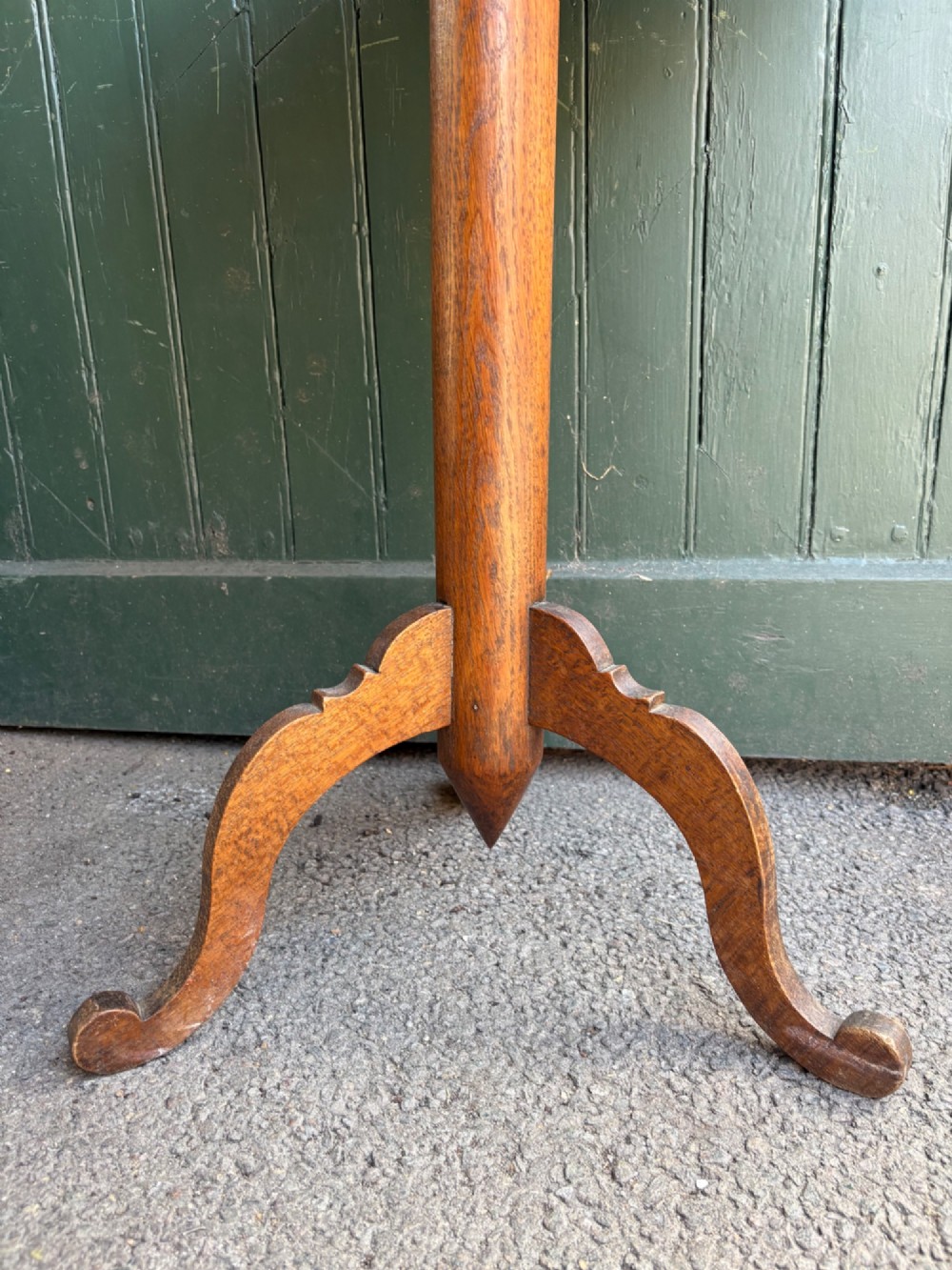 An English Arts and Crafts solid oak wine table featuring a circular top, an under-top support bracket, a central column terminating in a pointed lower finial, and three splayed cabriole legs. View V