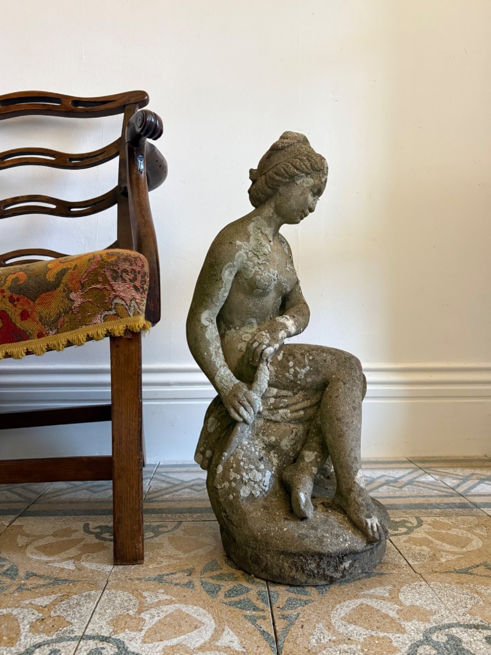 A weathered cast stone statue depicting a seated classical female bather figure with a natural lichen patina.