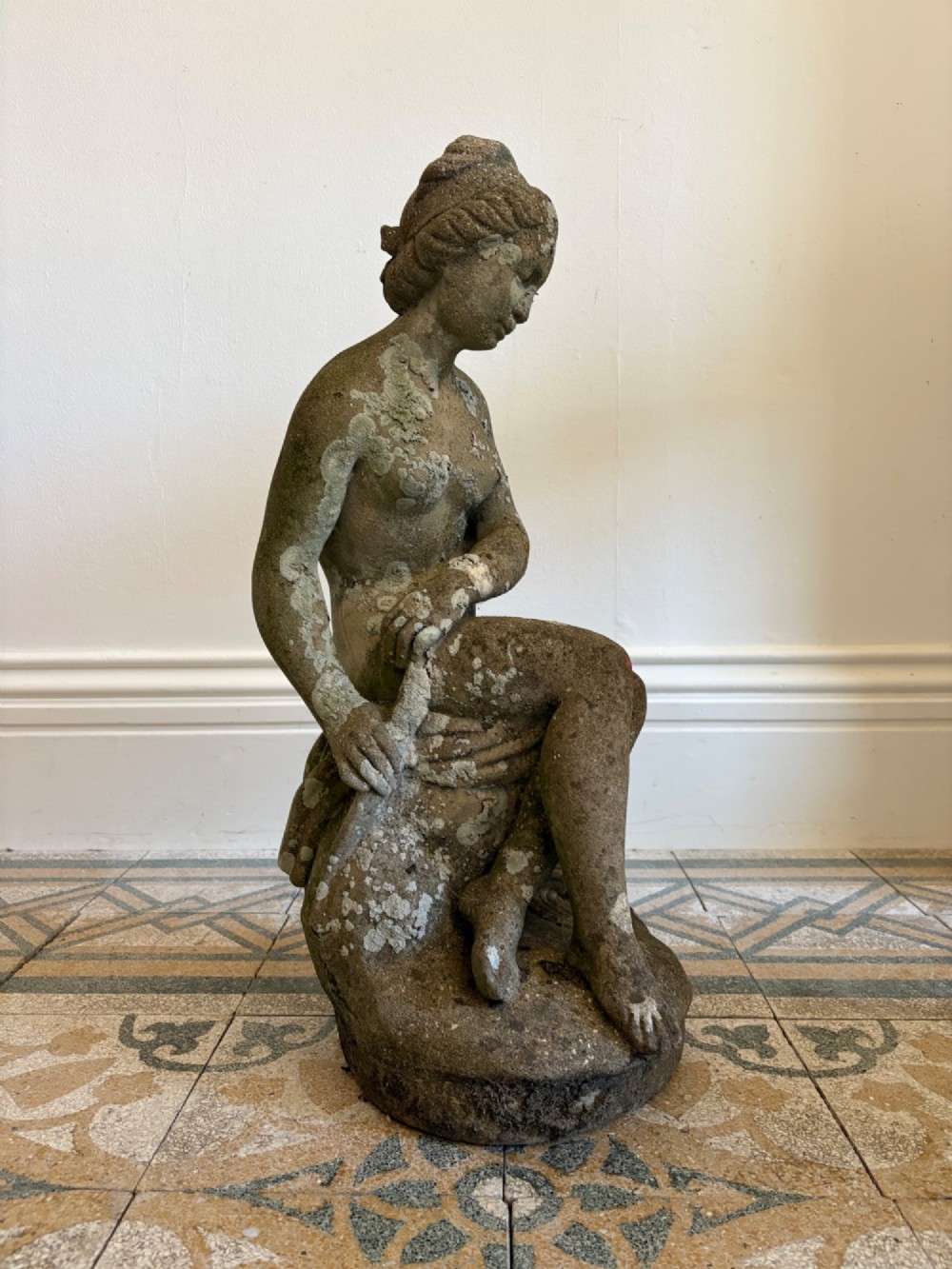 A weathered cast stone statue depicting a seated classical female bather figure with a natural lichen patina. View II