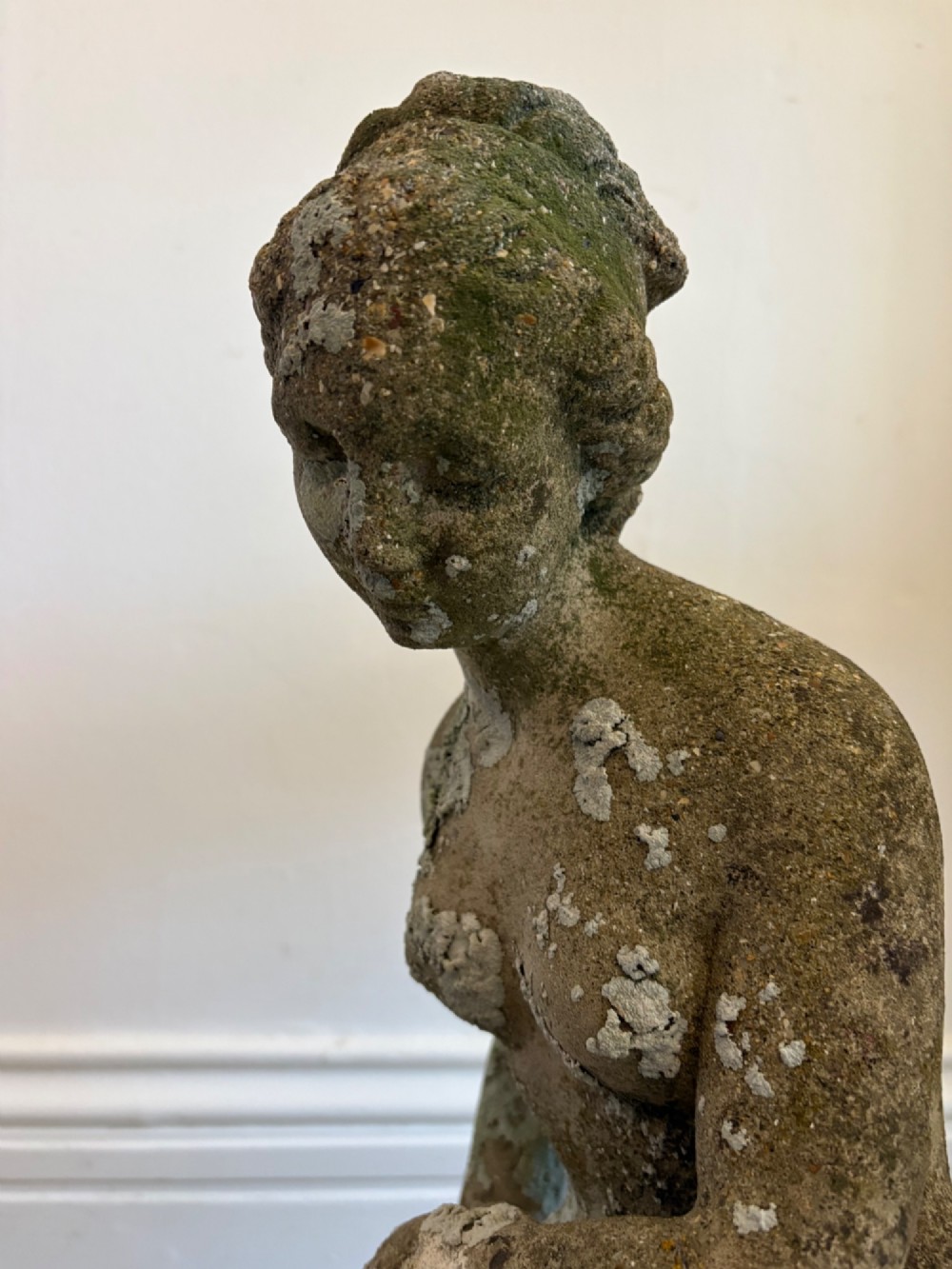 A weathered cast stone statue depicting a seated classical female bather figure with a natural lichen patina. View III