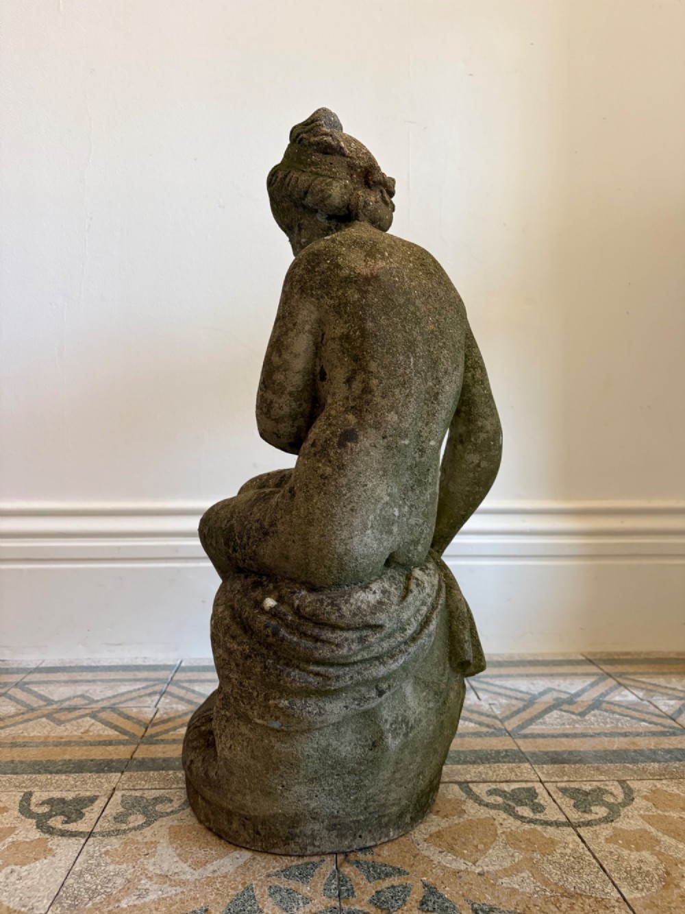 A weathered cast stone statue depicting a seated classical female bather figure with a natural lichen patina. View IV