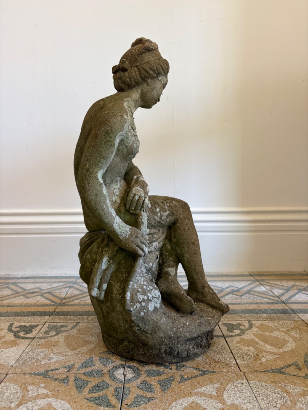 A weathered cast stone statue depicting a seated classical female bather figure with a natural lichen patina. View V