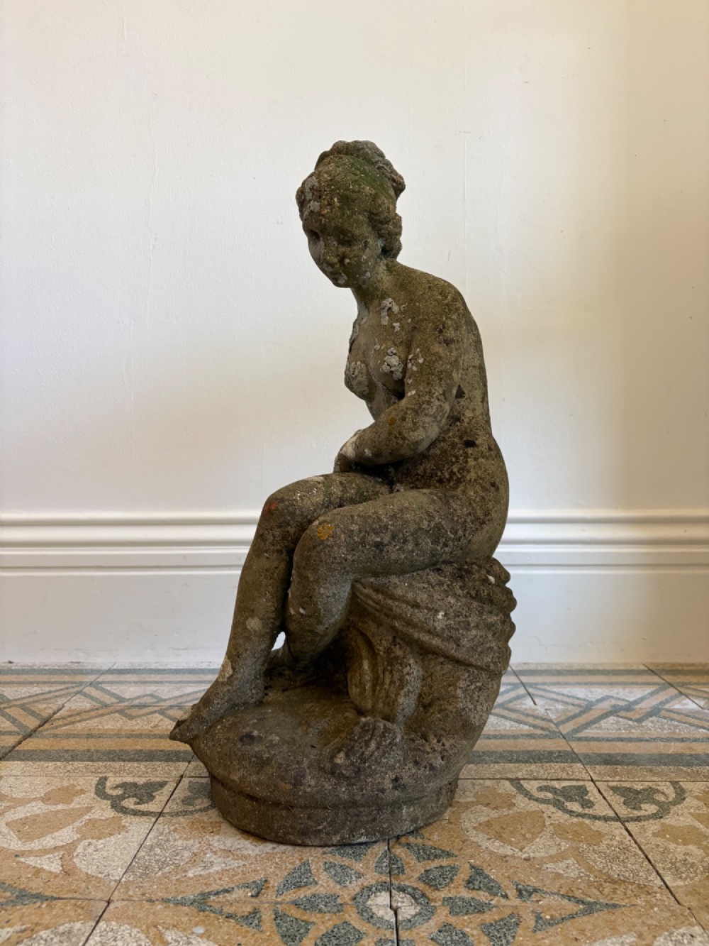 A weathered cast stone statue depicting a seated classical female bather figure with a natural lichen patina. View VI