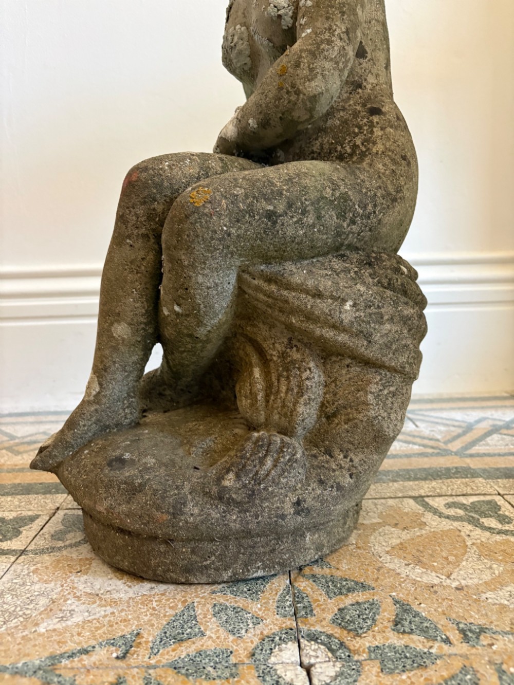 A weathered cast stone statue depicting a seated classical female bather figure with a natural lichen patina. View VII