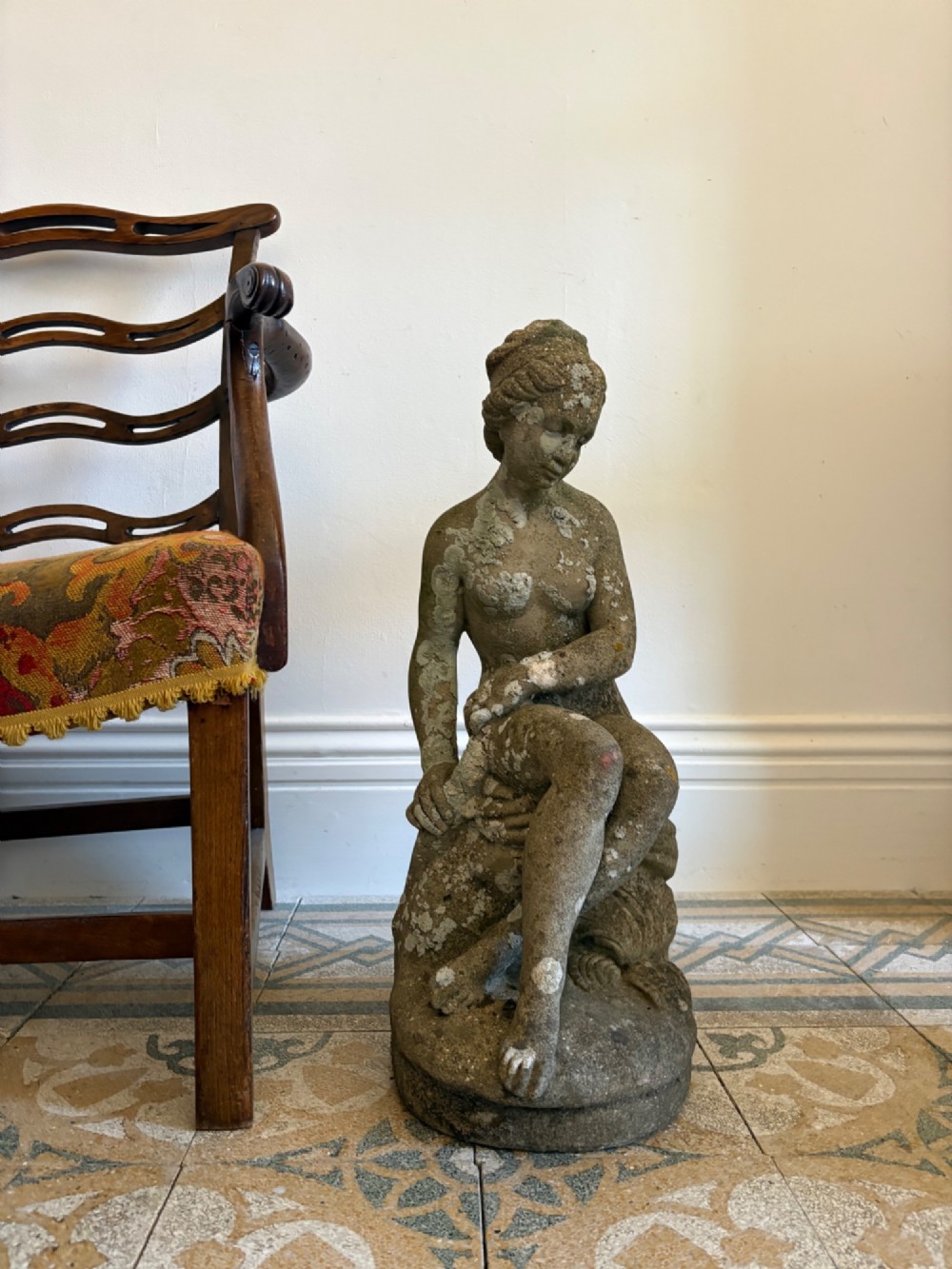 A weathered cast stone statue depicting a seated classical female bather figure with a natural lichen patina. View XIV