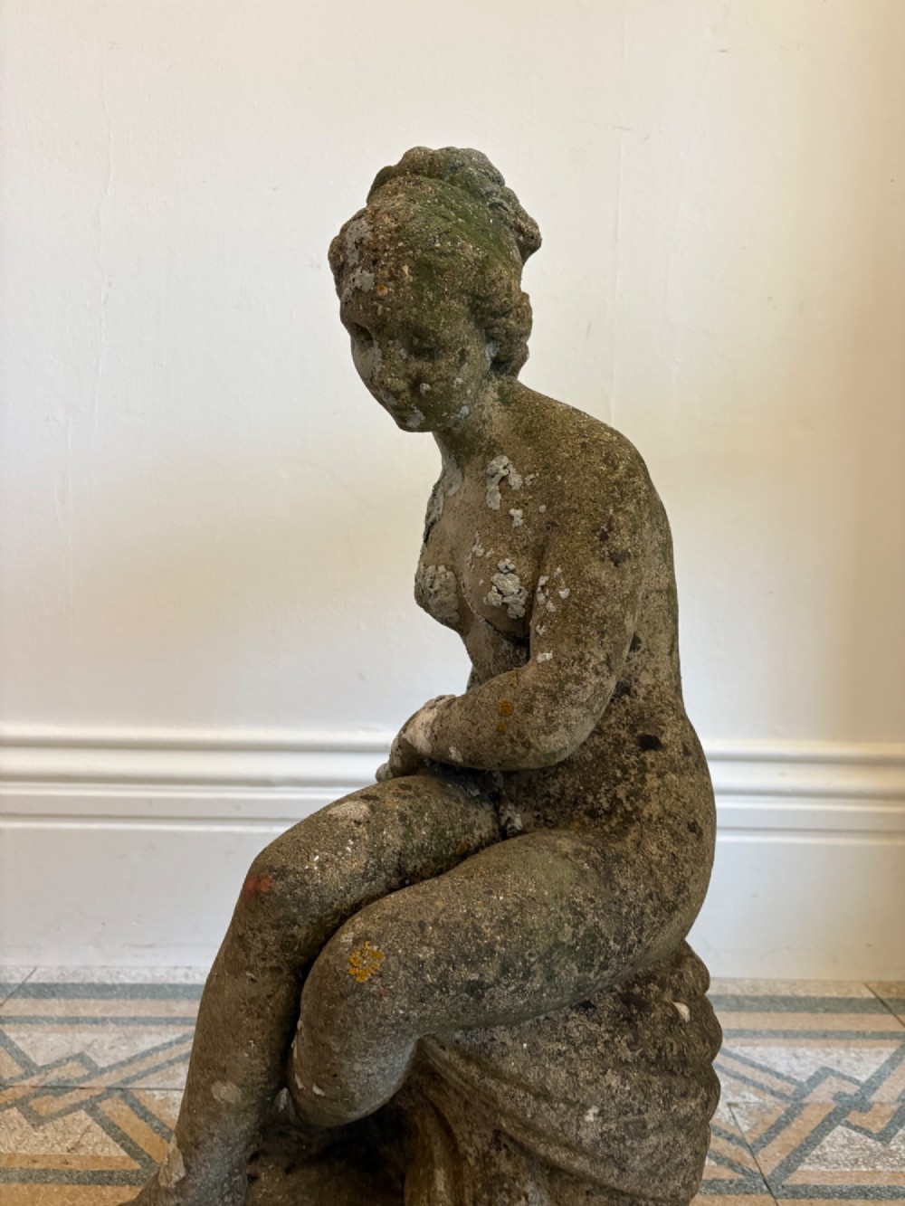A weathered cast stone statue depicting a seated classical female bather figure with a natural lichen patina. View XV