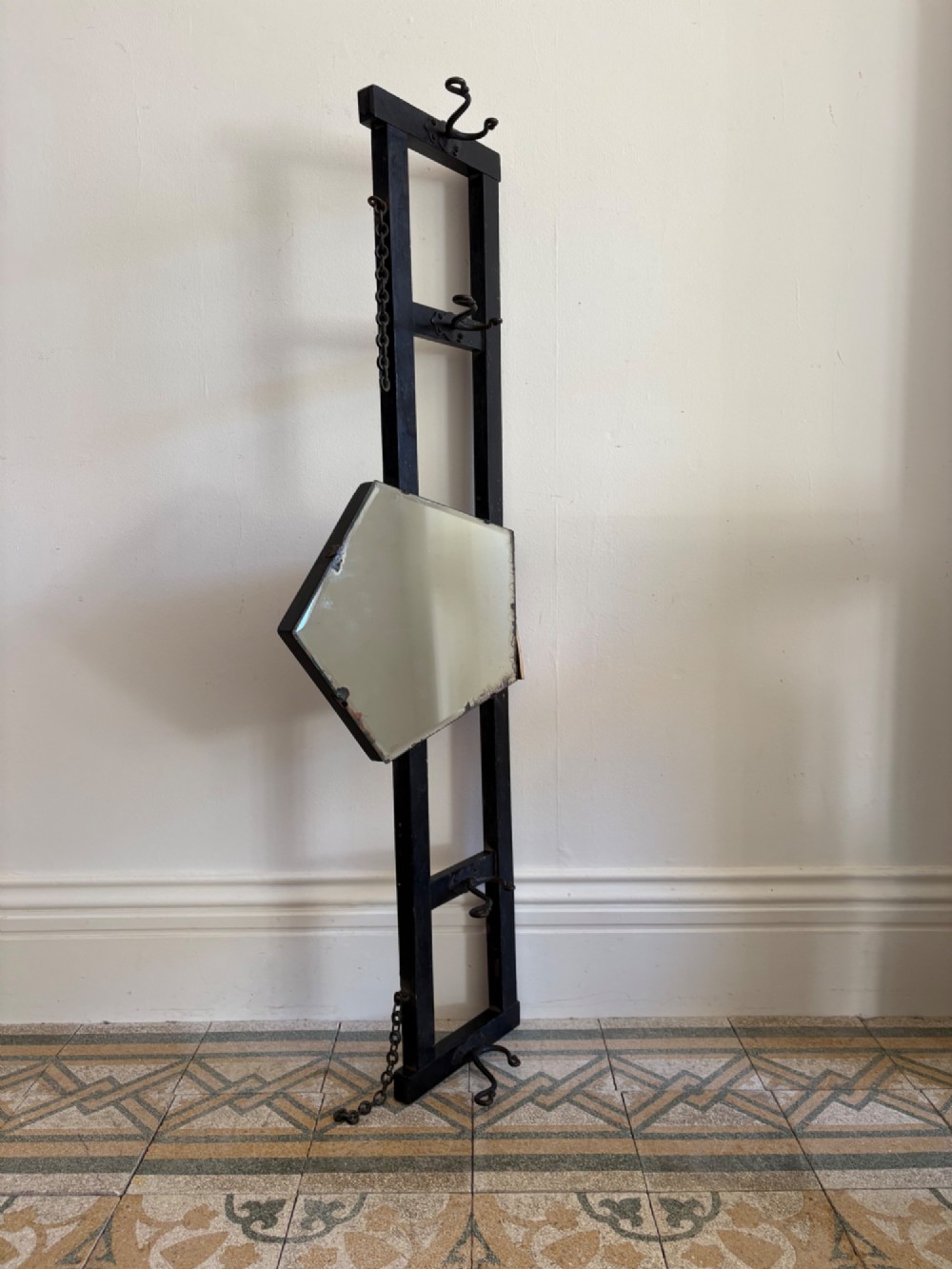 A tall black wrought iron wall-mounted coat rack featuring integrated hooks, hanging chains, and an asymmetrical pentagonal mirror.