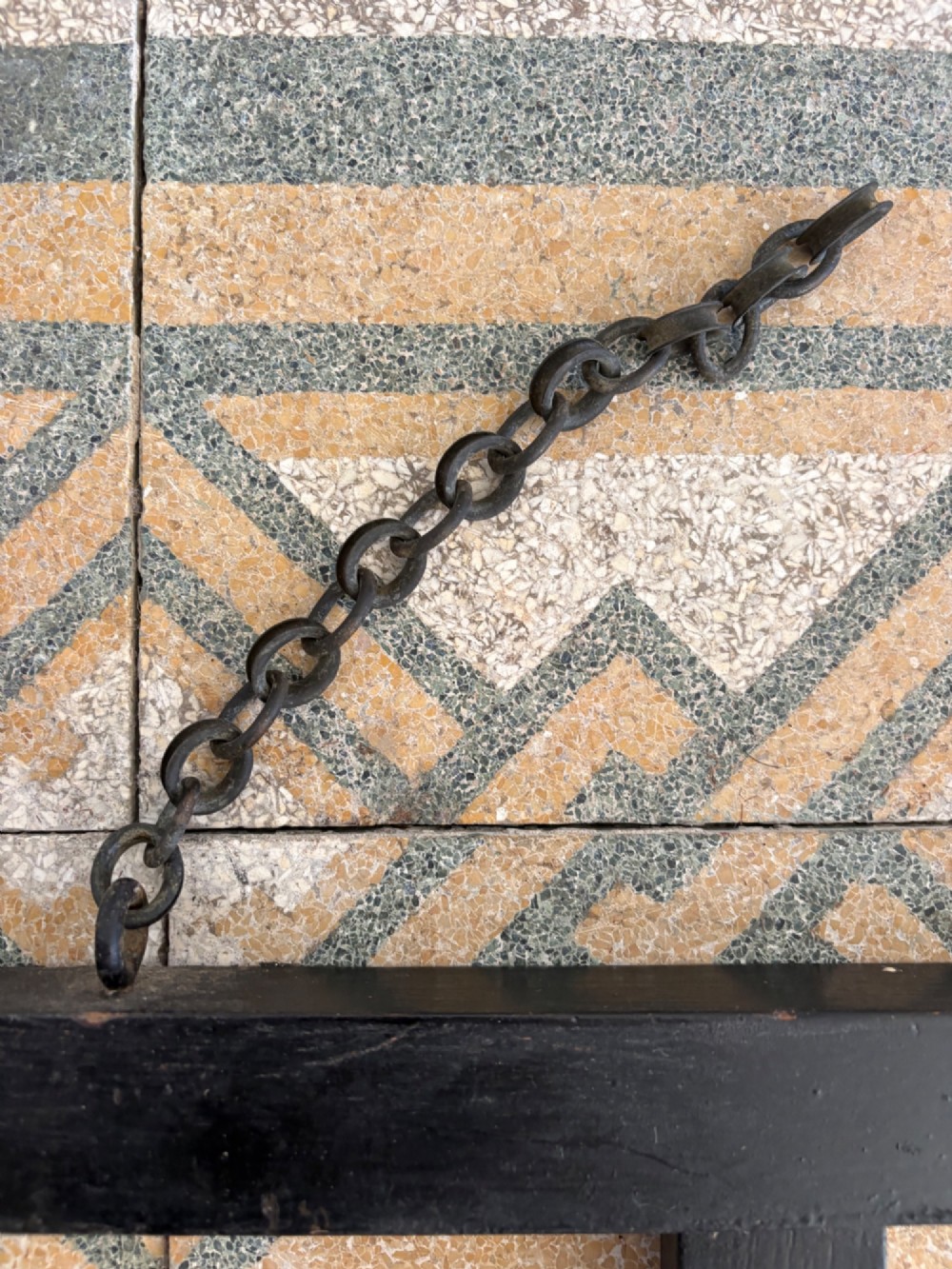 A tall black wrought iron wall-mounted coat rack featuring integrated hooks, hanging chains, and an asymmetrical pentagonal mirror. View VI