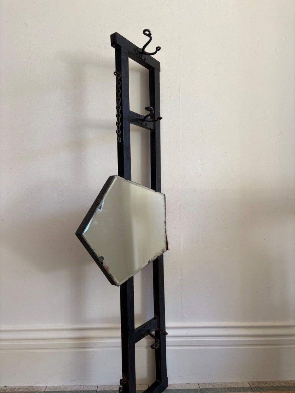 A tall black wrought iron wall-mounted coat rack featuring integrated hooks, hanging chains, and an asymmetrical pentagonal mirror. View VII