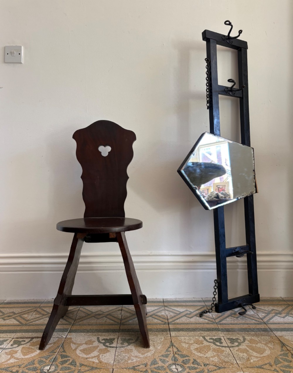 A tall black wrought iron wall-mounted coat rack featuring integrated hooks, hanging chains, and an asymmetrical pentagonal mirror. View X