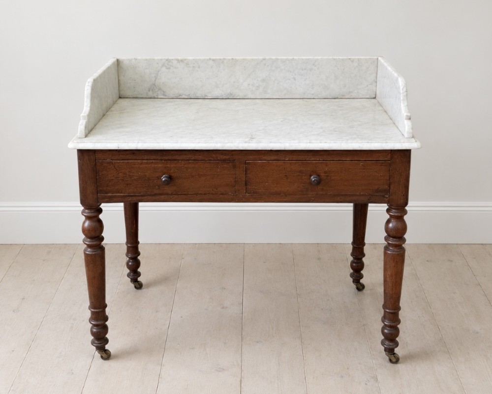 A 19th-century mahogany washstand featuring a white Carrara marble top with splashback, two frieze drawers with wooden knobs, and turned legs with brass castors.