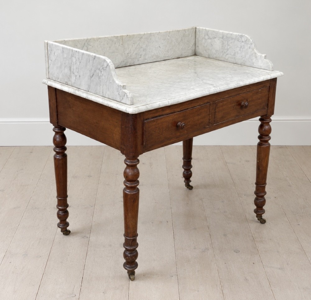 A 19th-century mahogany washstand featuring a white Carrara marble top with splashback, two frieze drawers with wooden knobs, and turned legs with brass castors. View II