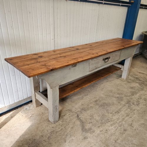 fig 2. A long rustic pine worktable featuring a natural wood top, a light grey painted frame with a single drawer, and a lower shelf.