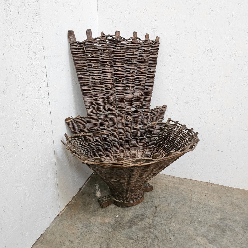 An antique French wicker grape harvester basket featuring a flared woven willow container resting on a wooden base with an extended vertical backrest. View II