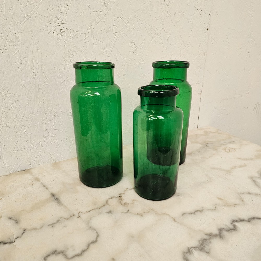 A set of three cylindrical green glass pickling jars of varying heights arranged on a light surface. View II
