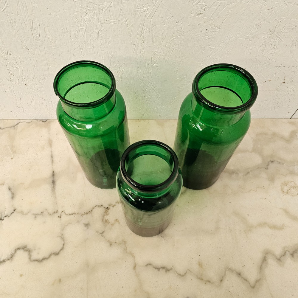 A set of three cylindrical green glass pickling jars of varying heights arranged on a light surface. View III