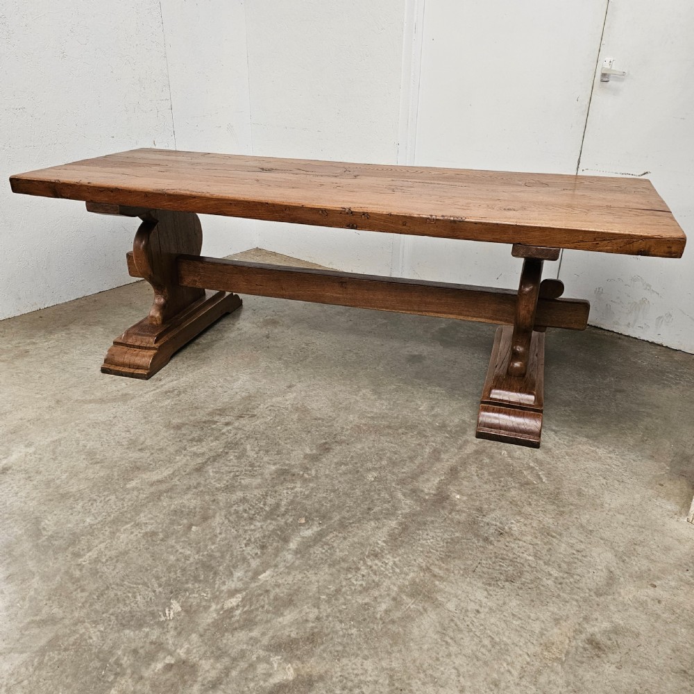 A long French pale oak trestle dining table featuring a solid plank top supported by shaped pedestal legs and a low central stretcher.